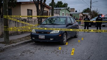 Un automóvil azul acordonado con cinta amarilla de la policía estatal y casquillos de bala junto a marcadores de evidencia en la calle.
