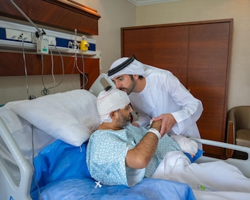Un hombre con vestimenta tradicional blanca besa la cabeza vendada de un paciente acostado en una cama de hospital mientras sostiene su mano