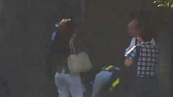 Varias personas caminan por una calle junto a una pared, entre ellas una mujer con bolso blanco y un hombre con camisa a cuadros