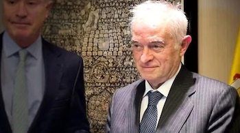 Un hombre de cabello blanco con traje rayado y corbata de puntos. Otro hombre de traje oscuro. Una pared con tapiz y una bandera amarilla y roja.