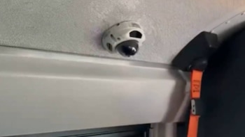 Una cámara de seguridad domo blanca y negra está montada en un techo de color claro. Abajo hay un panel blanco y a la derecha una correa naranja colgando