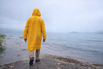 Pessoas por trás, vestindo capa de chuva amarela e calças cáqui, caminhando na margem do lago ou no oceano sob chuva fraca.