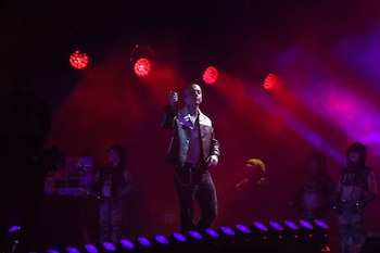 Un cantante sobre una tarima con focos de luz roja y humo, rodeado por bailarines con ropa oscura