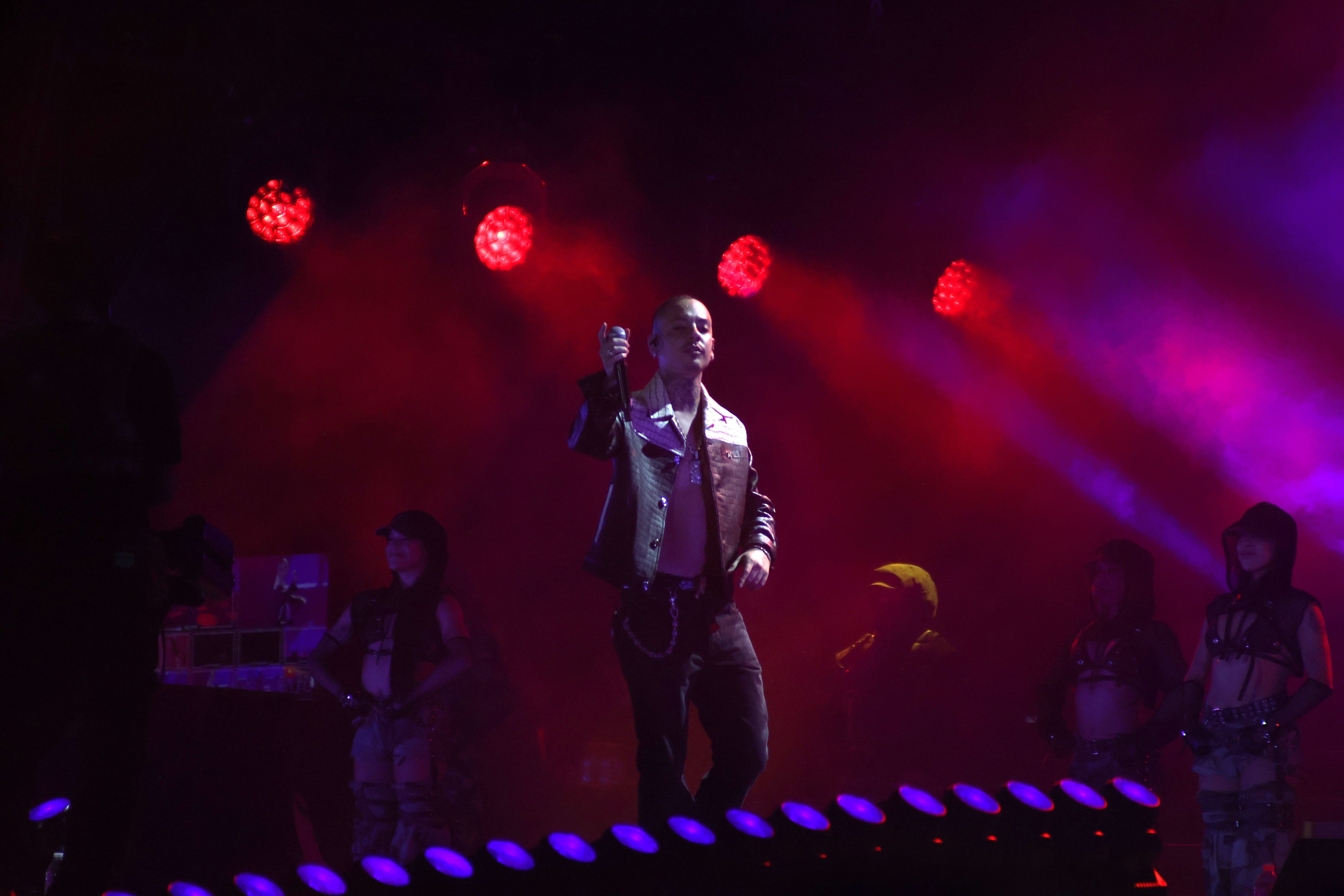 Un cantante sostiene un micrófono sobre un escenario iluminado por luces rojas durante un concierto en vivo. (Selene Miranda/Infobae México)