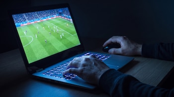 Una laptop abierta muestra un videojuego de fútbol. Dos manos manipulan el teclado retroiluminado y un ratón sobre una mesa de madera en la oscuridad.