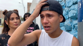 Primer plano de Sebastián Gálvez llorando, con una mano en la cabeza, junto a una mujer. Detrás, Valentino observa. Fondo de calle con muro azul texturizado.