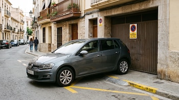 Automóvil gris aparcado junto a la acera frente a un portón con una placa de vado permanente y líneas amarillas en la calzada.