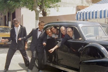 Hombres vestidos con trajes oscuros posan junto a un automóvil antiguo de color negro en una calle