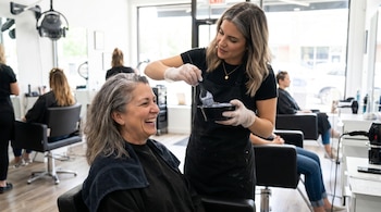 Estilista aplica tinte a las raíces del cabello gris de una mujer sonriente. Ambas están en un salón de belleza, con otros clientes de fondo.