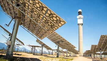 Campo con numerosas estructuras de paneles solares sobre postes metálicos y una alta torre gris con la parte superior negra y blanca, bajo cielo azul