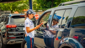 Una mujer de uniforme blanco y gorra azul sostiene un dispositivo rojo y habla con un conductor dentro de un SUV. Un segundo SUV negro está detrás. Hay un cartel de alto