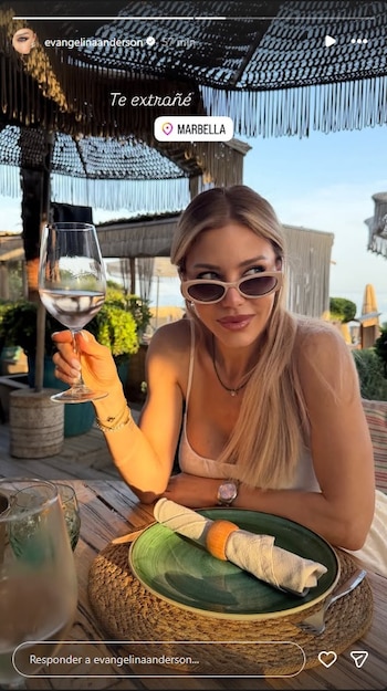 Mujer rubia con gafas de sol y vestido blanco sentada en una mesa de madera, sostiene una copa con líquido. Hay un plato con una servilleta