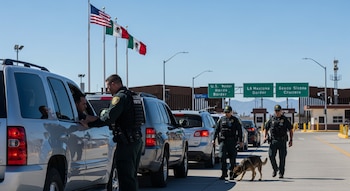 Varios agentes de aduanas inspeccionan vehículos en un punto fronterizo entre Estados Unidos y México, con perros detectores y banderas de ambos países visibles.