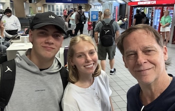 Selfie de tres personas sonrientes en el frente. Un hombre adulto, una mujer joven y un hombre joven con gorra. Fondo concurrido de aeropuerto