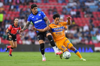 Dos jugadores de fútbol, uno con camiseta azul, el otro con camiseta naranja y azul, disputan un balón en un campo de césped. Una árbitra de rojo de fondo