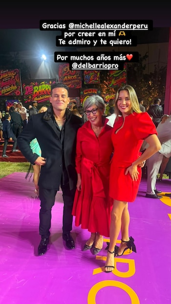 Un hombre, una mujer mayor con gafas y una mujer joven posan sonrientes en una alfombra morada. Hay carteles de fondo con la leyenda 'Del Barrio Producciones'