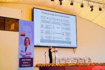 Una ponente habla junto a un podio frente a una pantalla de presentación y un cartel vertical con el nombre de la doctora Maria J. Rodríguez