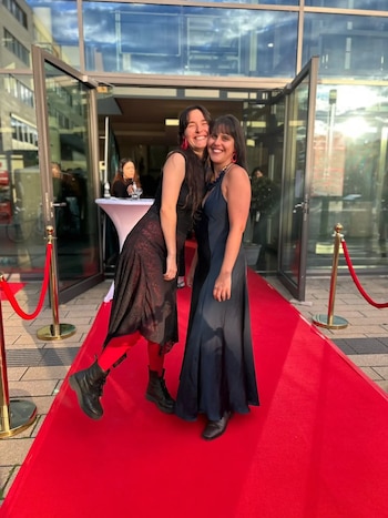 Dos mujeres sonríen sobre una alfombra roja. La mujer de la izquierda lleva falda negra, medias rojas y botas; la de la derecha, vestido azul largo. Edificio de cristal al fondo