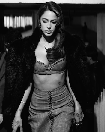 Mujer con cabello oscuro, chaqueta de piel, bralette de tartán con cordones, falda de tartán, pulseras y tatuajes en las manos