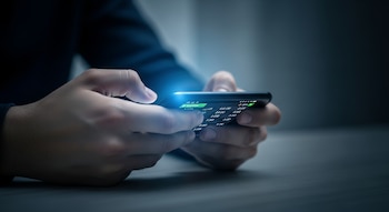 Manos masculinas sostienen un teléfono inteligente con pantalla iluminada en la oscuridad, mostrando una interfaz borrosa sobre una mesa.