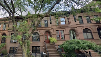 Fachada de una hilera de edificios residenciales de tres pisos con ladrillos marrones, escaleras de entrada, puertas arqueadas y árboles frondosos en primer plano