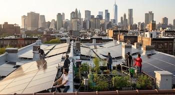 Varias personas en azoteas de Nueva York, algunas instalando paneles solares y otras cuidando jardines. El horizonte urbano con rascacielos se ve al fondo.