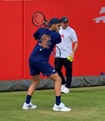 Francisco Cerúndolo golpea una pelota de tenis con raqueta en una cancha de césped. Viste ropa deportiva oscura, gorra azul, con una pared roja y edificios al fondo
