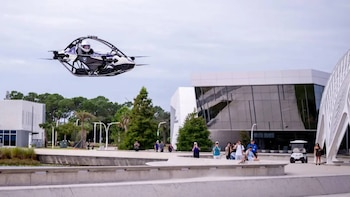 Una aeronave monoplaza tripulada vuela a baja altura sobre una plaza peatonal frente a edificios modernos y un grupo de personas
