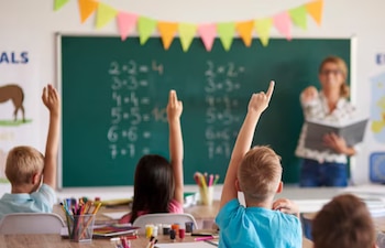 Niños sentados en pupitres en un aula con las manos levantadas. Una maestra borrosa apunta a un alumno. Hay una pizarra verde con ecuaciones y guirnaldas
