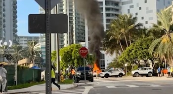 Una calle urbana con un vehículo en llamas, una densa columna de humo negro, patrullas policiales y edificios al fondo