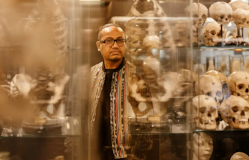 Hombre con gafas y chaqueta multicolor de pie frente a vitrinas de vidrio que exhiben múltiples cráneos y restos óseos humanos