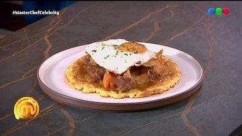 Un plato blanco con una base redonda de masa, trozos de carne con verduras y un huevo frito encima, dispuesto sobre una mesa oscura