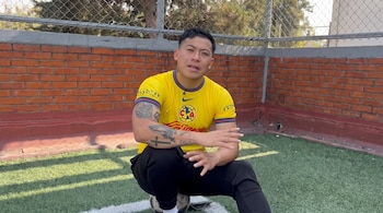 Un hombre con tatuajes y camiseta amarilla del Club América se sienta sobre un balón en una cancha sintética con pared de ladrillo.