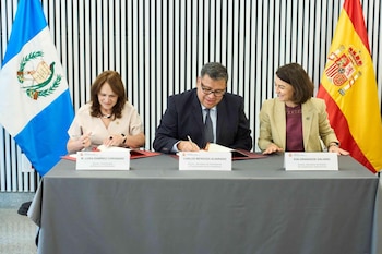Tres personas sentadas en una mesa firman documentos. Hay dos banderas de países, una de Guatemala y una de España. Tres carteles de nombres se ubican frente a cada persona