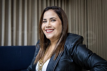 Mujer con cabello castaño largo sonriendo, usa chaqueta de cuero negra, blusa blanca y múltiples collares dorados, sentada en sofá azul oscuro, fondo de cortina beige