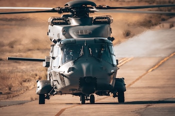 Helicóptero militar de color verde grisáceo con rotores principales y de cola, dos pilotos en la cabina, avanzando sobre una pista de asfalto, con estela de humo