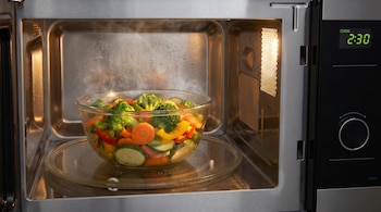 Verduras picadas (brócoli, zanahorias, pimientos, calabacín) en un tazón de vidrio se cocinan con vapor dentro de un microondas con la luz encendida.