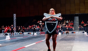 Una mujer sostiene un escudo metálico grande sobre el pecho mientras camina en una arena con público al fondo