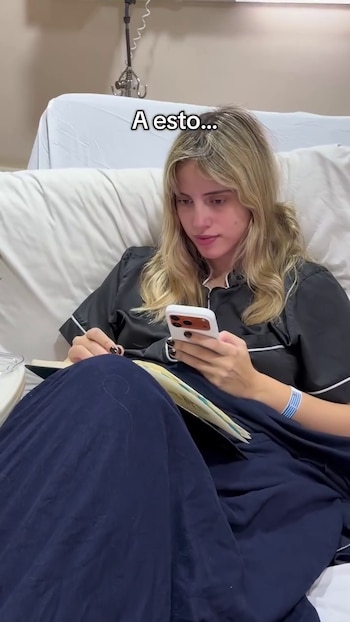 Mujer joven rubia en cama de hospital, vestida con pijama negro, usa su teléfono celular con una mano y sostiene un libro con la otra, tiene una pulsera de internación