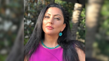 Primer plano de una mujer con cabello oscuro largo, top magenta y joyas turquesas, mirando a la cámara con luz solar en su rostro y fondo borroso