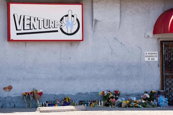 Un cartel de Venture N sobre una pared de textura gris con ramos de flores, velas encendidas y marcos de fotos en el suelo