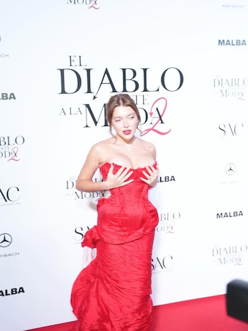 Sofie la Reini Gpnet, con vestido rojo strapless y collar, posa en una alfombra roja. El fondo blanco muestra el logo 'El diablo viste a la moda 2'
