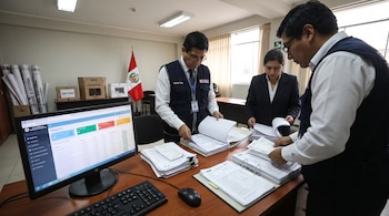 Tres personas revisan documentos en un escritorio con computadora. Fondo: bandera de Perú, urnas de votación y rollos con inscripciones electorales