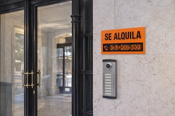 Cartel naranja con texto 'Se alquila' y número de teléfono pixelado sobre un telefonillo en una pared de mármol blanco, junto a una puerta negra de cristal.