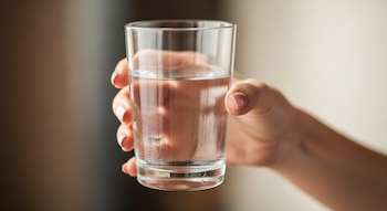 Una mano femenina sostiene un vaso de vidrio transparente con agua. El vaso presenta gotas de condensación en su superficie externa.