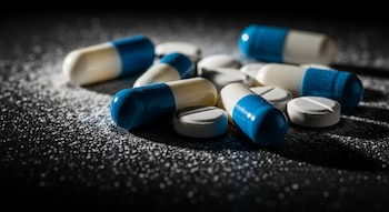 Primer plano de varias pastillas: algunas cápsulas azules y blancas, y algunas tabletas redondas blancas, esparcidas sobre una superficie oscura con polvo blanco.