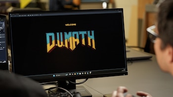 Primer plano de una pantalla de computadora con el título "DU.MATH" en fuente estilizada; una persona con gafas y las manos en un controlador de juego está viendo la pantalla