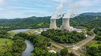 Vista aérea de una planta nuclear con dos torres de enfriamiento emitiendo vapor blanco, paneles solares, un río y colinas verdes en El Salvador bajo un cielo nublado.