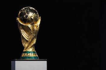 Vista frontal del Trofeo de la Copa Mundial de la FIFA dorado y verde sobre un pedestal blanco, con un fondo negro liso. El trofeo se sitúa a la izquierda.