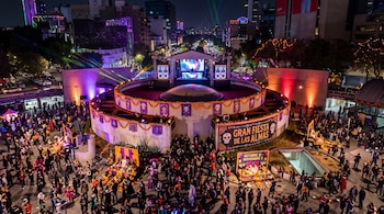 Plaza pública de noche con personas reunidas, altares tradicionales, flores de cempasúchil y un escenario con pantallas y luces.